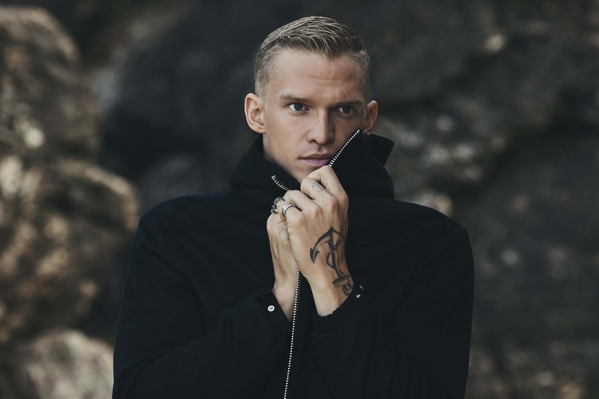 Portrait of a blond adult male in a black jacket against a blurred background. The man has a tattoo on his hand.