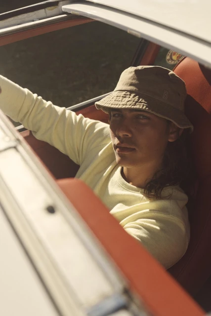 Young adult with a tan bucket hat and light yellow sweater sitting in a red car, looking at camera. Outdoor setting with natural light.