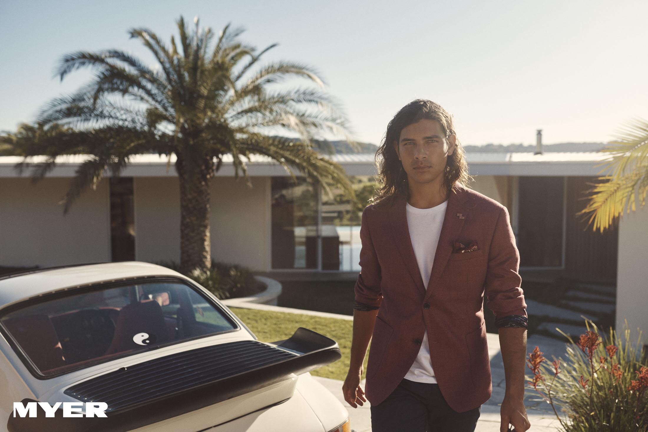 Man in burgundy blazer with car. Stylish male fashion model poses outdoors next to a vintage white Porsche car against a modern home.