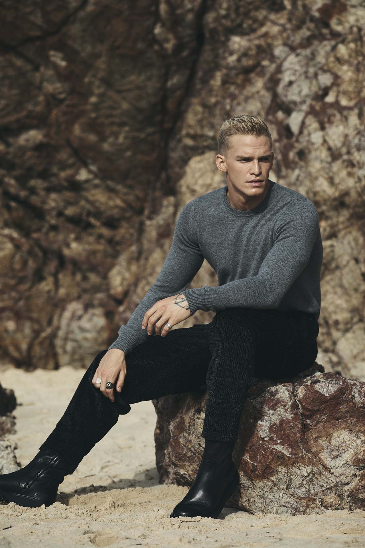 Male fashion model in gray sweater poses on a rock in an outdoor setting. He wears black pants and boots against a rocky background.