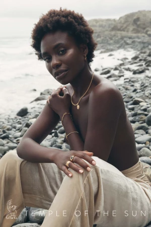 Black woman wearing gold jewellery sitting on a rocky coast with light-coloured trousers.
