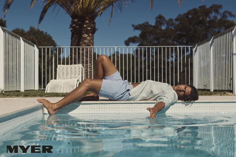 A man lying on the edge of a swimming pool with a palm tree in the background on a sunny day, wearing shorts and a shirt.