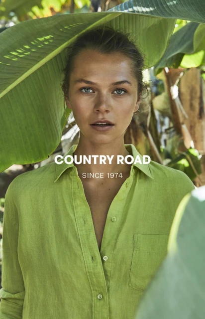 Close-up portrait of a woman in a green button-down shirt surrounded by large green leaves, showcasing a fashion or beauty aesthetic.