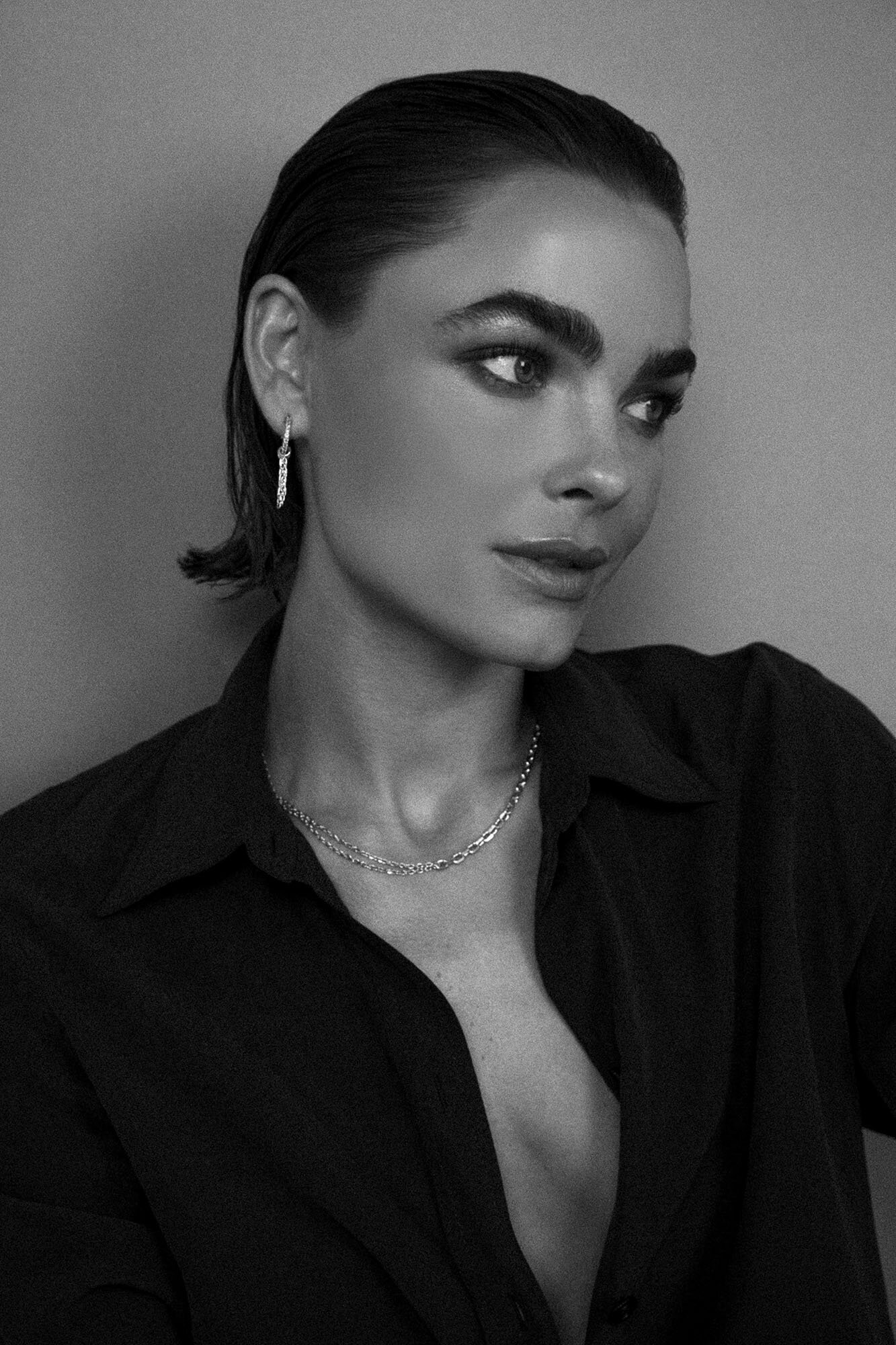 Monochrome portrait of a woman with slicked-back hair, wearing earrings and a partially unbuttoned black shirt with a silver necklace.