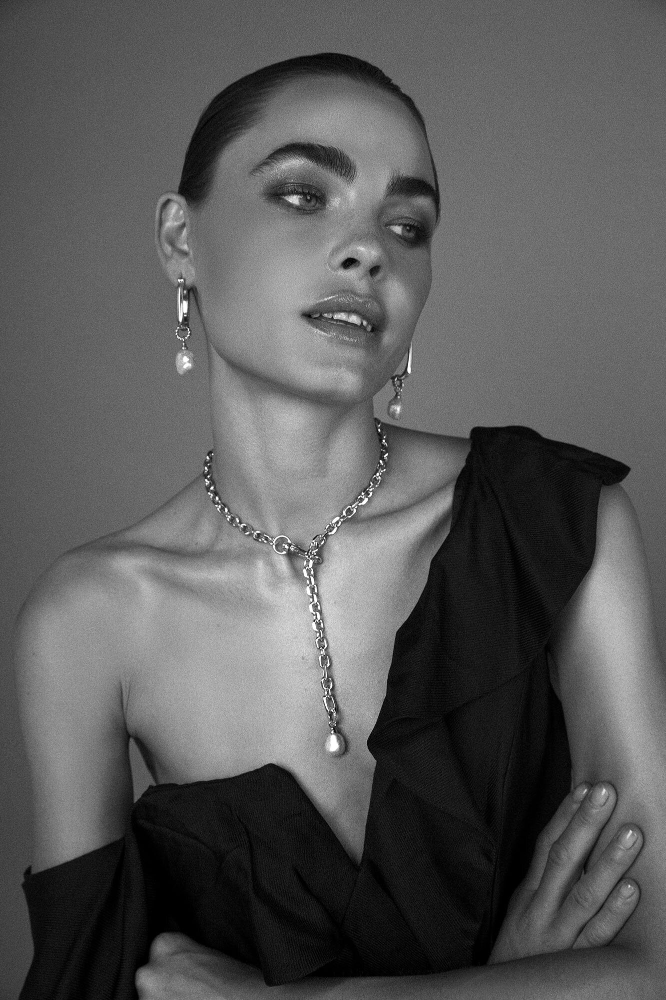 Black and white portrait of a young woman with dark hair, statement jewelry, and black ruffled top against a gray studio backdrop.