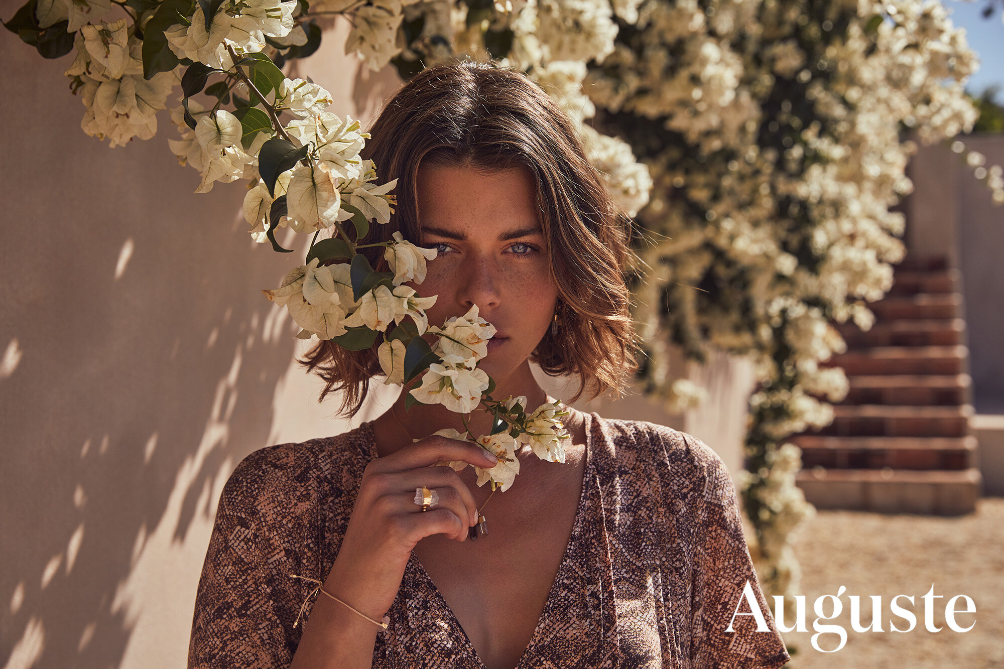 Portrait of a woman with short brown hair posing with white flowers outside in the daylight wearing a snake print dress.