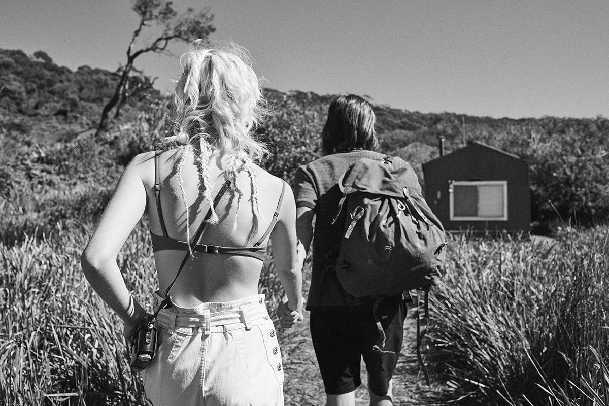 Two people hiking, viewed from behind. A woman with blonde braids and denim shorts holds hands with a backpacker on a trail.