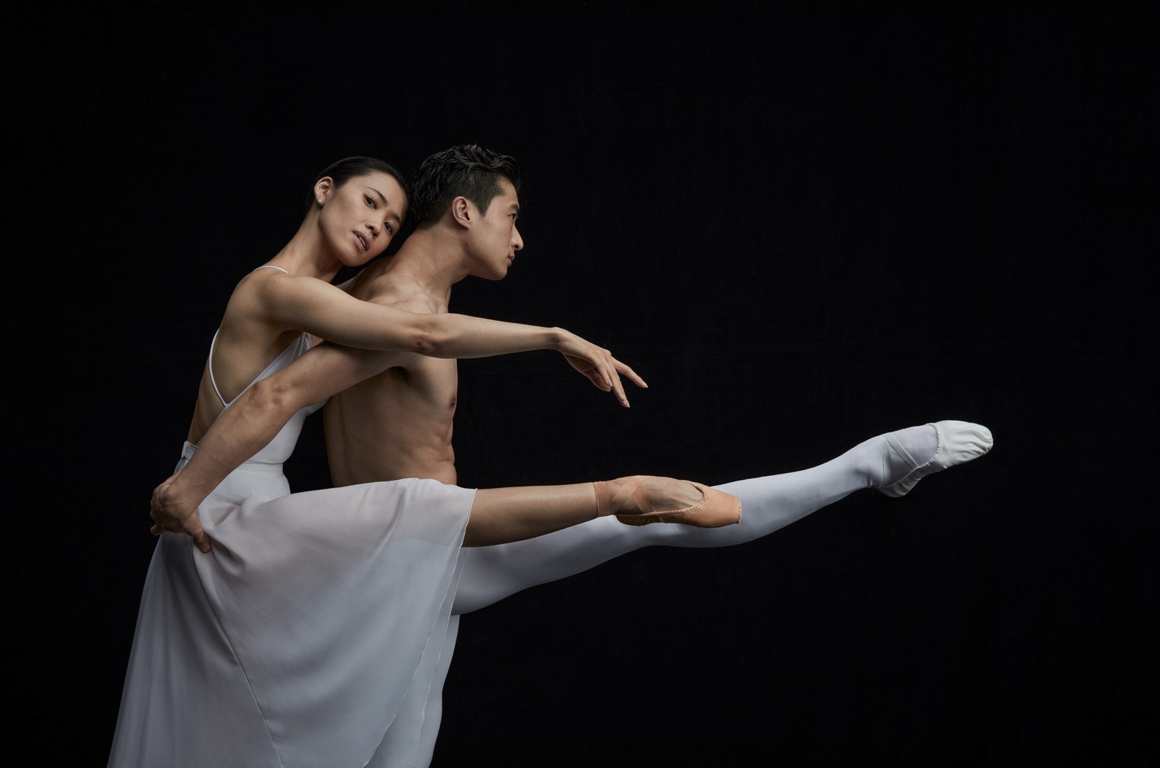 Ballet dancers performing together in a studio against a dark background. One dancer is leaning against the other with outstretched arms.