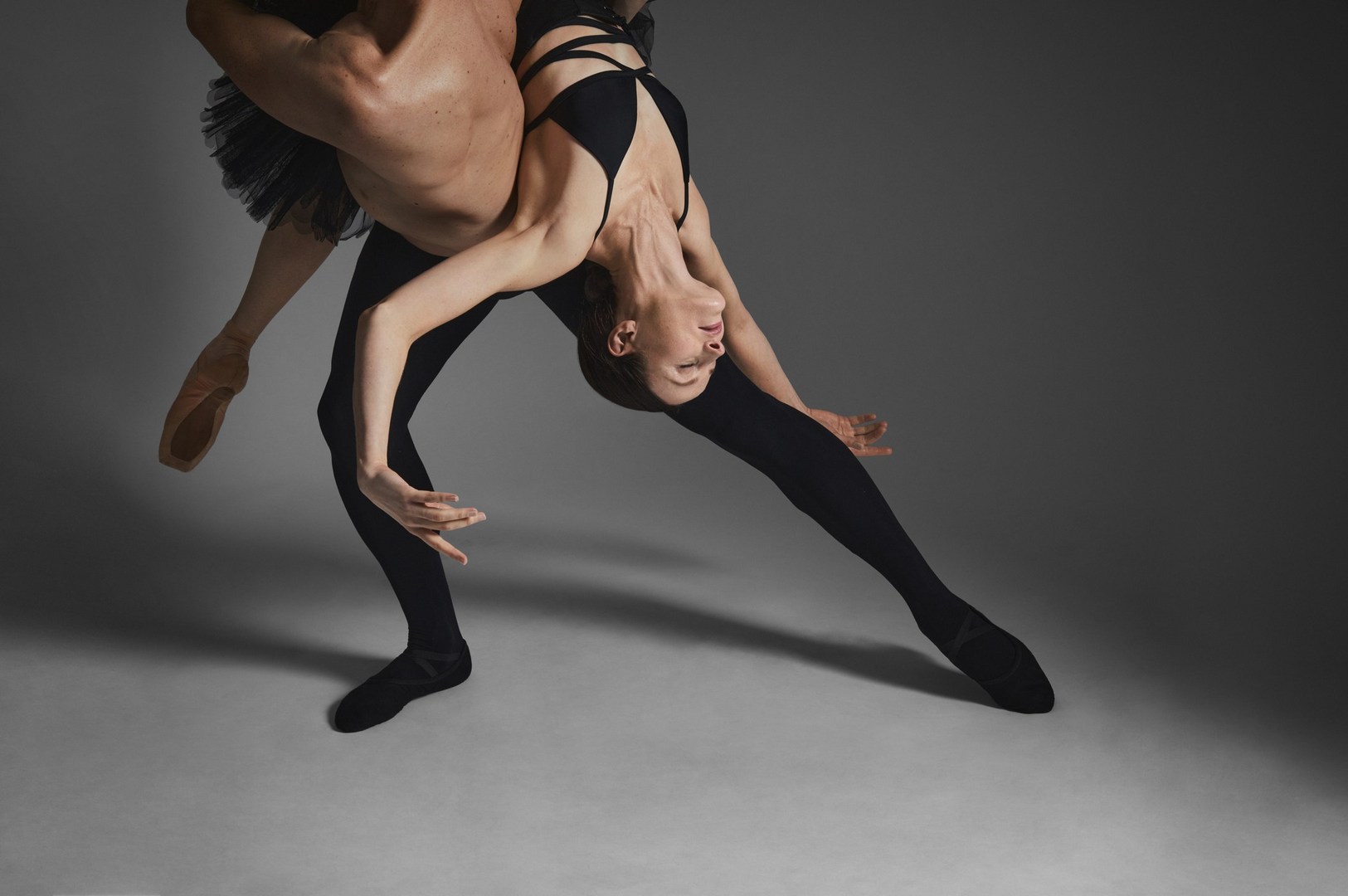 A male ballet dancer holds up a female ballet dancer in an artistic, sensual dance pose against a gray backdrop in studio lighting.