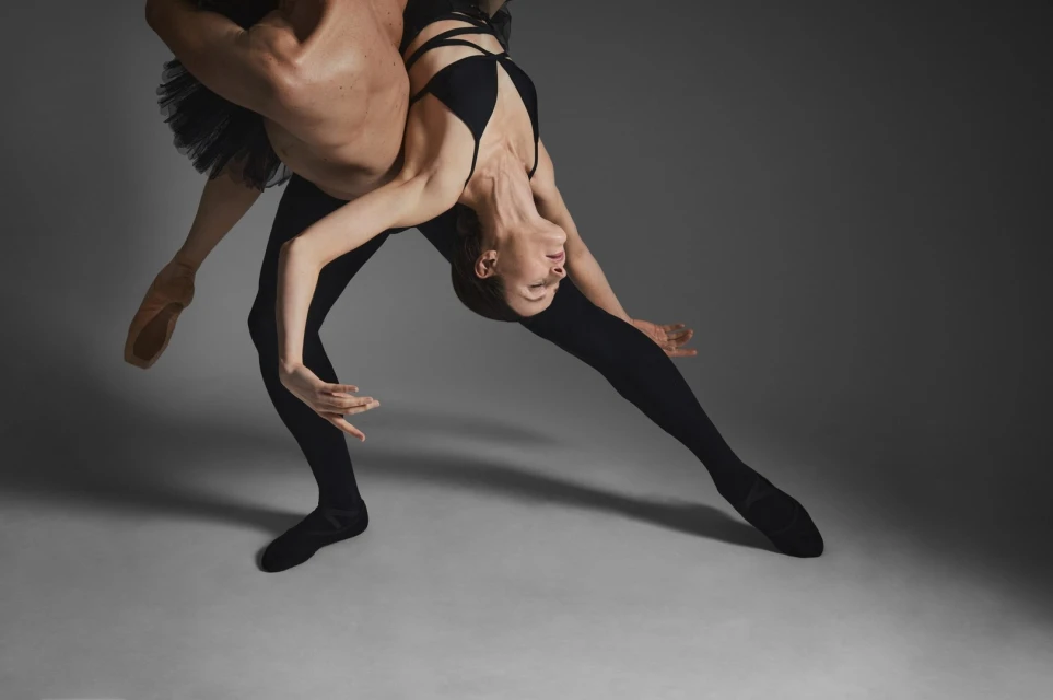 A male ballet dancer holds up a female ballet dancer in an artistic, sensual dance pose against a gray backdrop in studio lighting.