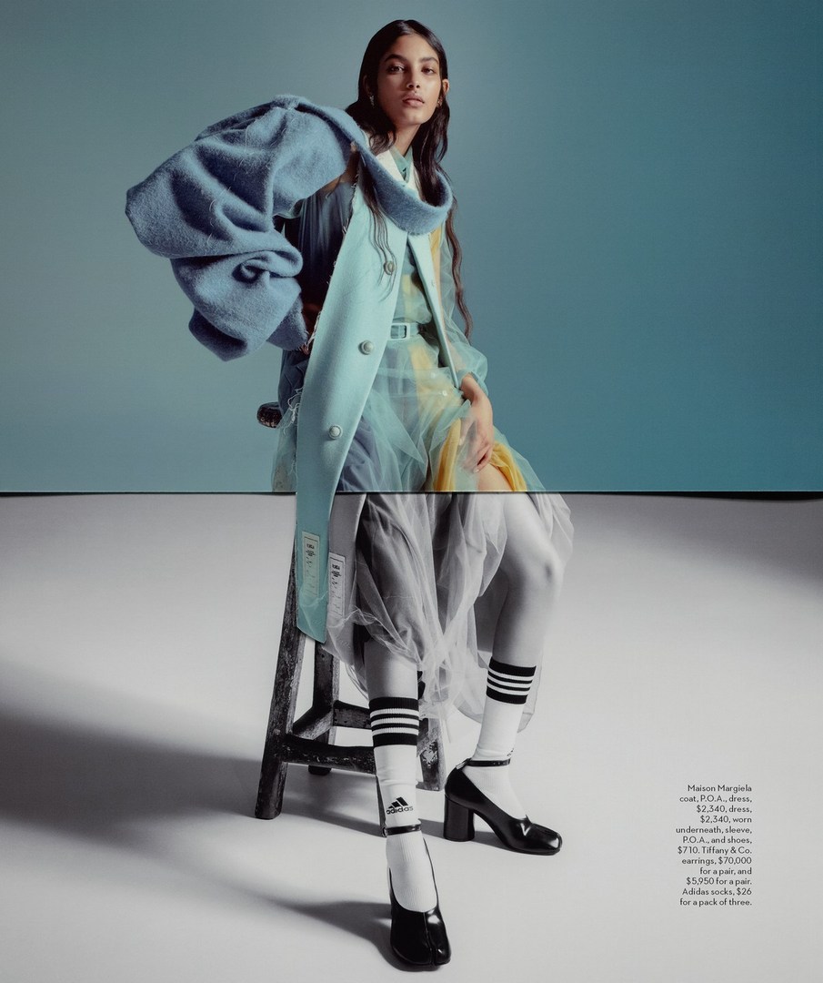 Fashion editorial shot of a woman in a pale blue coat, yellow dress, and Adidas socks sitting on a vintage wooden stool against a light blue backdrop.