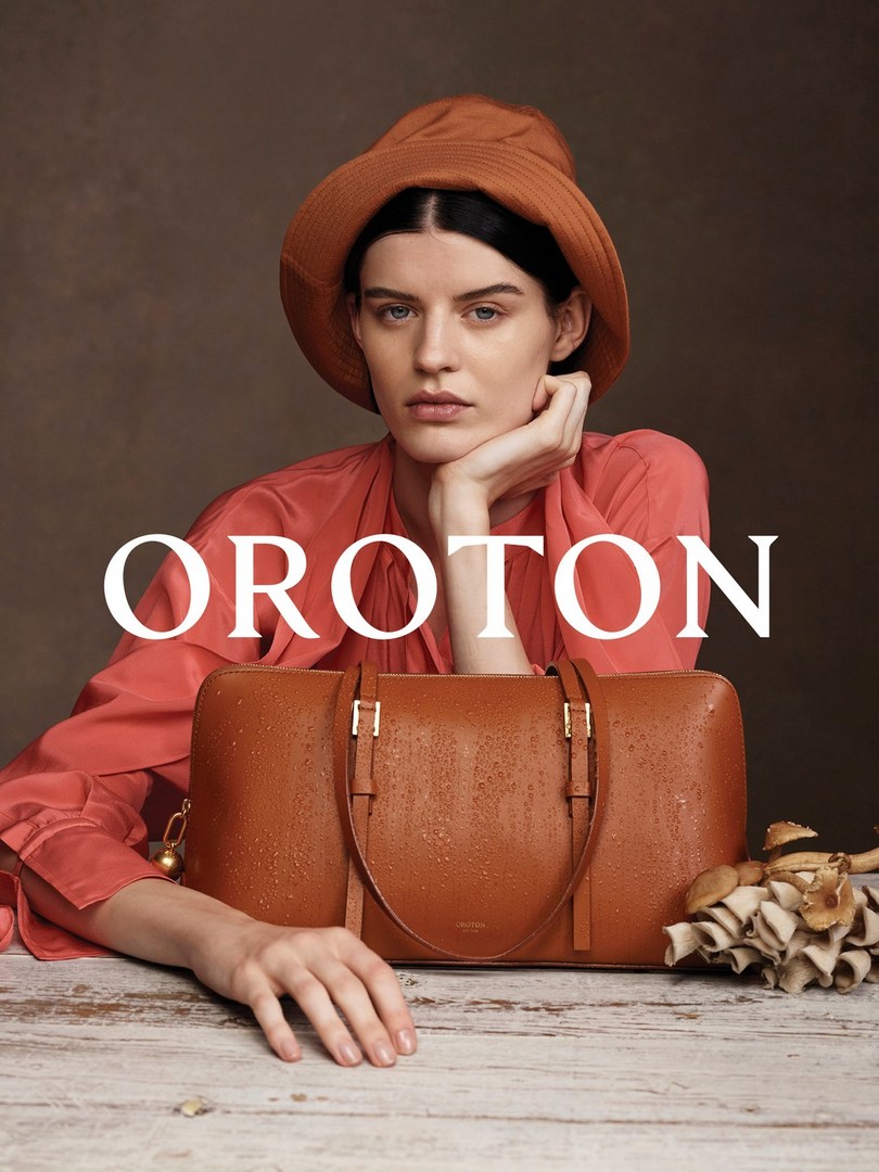 Woman wearing a hat poses with a brown leather handbag on a wooden surface, against a muted brown backdrop in a studio setting.