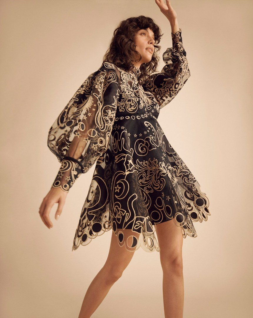 Woman in a black and gold patterned dress poses in a studio. The dress features puffed sleeves and a scalloped hemline.