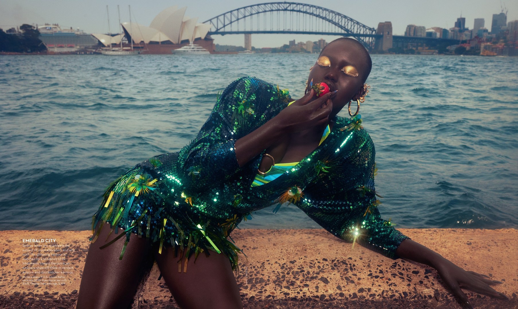 Adult female model in an emerald green sequin dress eating a strawberry near water with buildings and a bridge in the background.