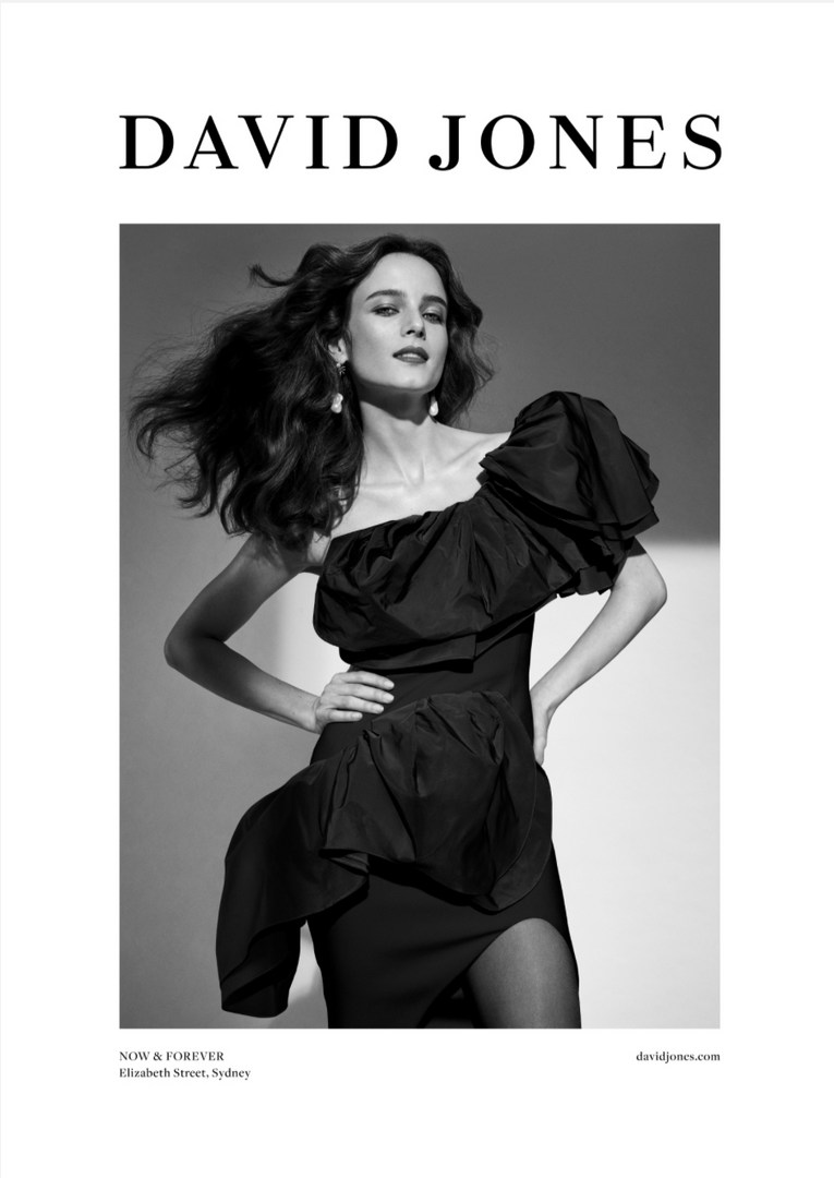 Fashion editorial shot of a woman in a black one-shoulder ruffle dress, wind-swept hair, and jewelry posing in a photo studio.