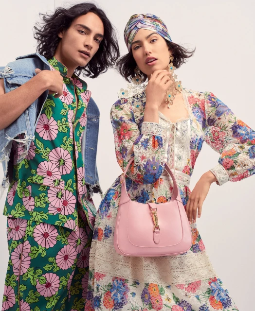 Two fashion models pose in floral print dresses: one in green with a denim jacket, the other in white with a pink handbag.