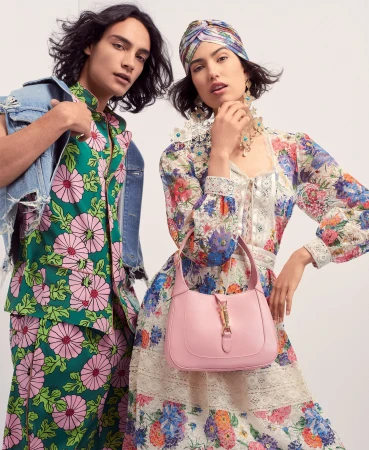 Two fashion models pose in floral print dresses: one in green with a denim jacket, the other in white with a pink handbag.