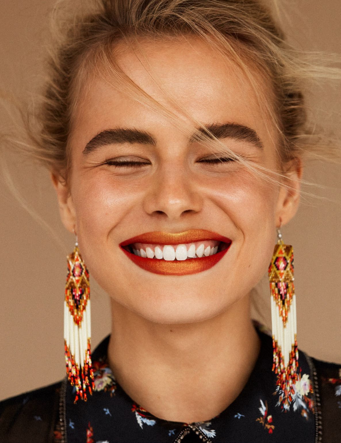 Close-up studio shot of a smiling woman with closed eyes, fair skin, and orange lipstick wearing beaded fringe earrings.