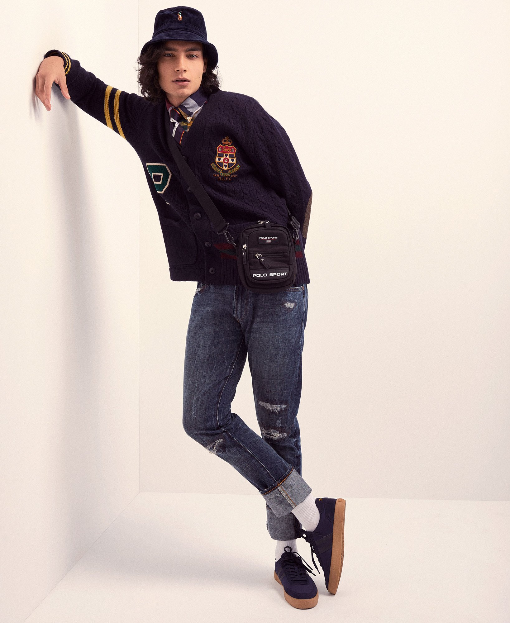A young adult male in a casual pose wearing a blue patterned bucket hat, cardigan, jeans, crossbody bag, and sneakers against a white wall.