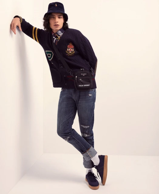 A young adult male in a casual pose wearing a blue patterned bucket hat, cardigan, jeans, crossbody bag, and sneakers against a white wall.