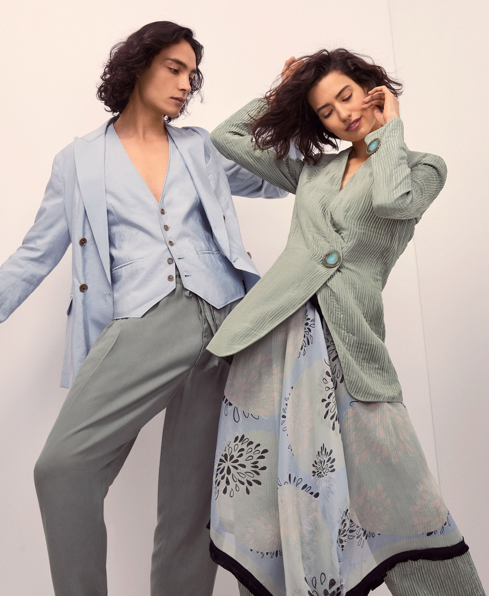 Two fashion models in a studio pose. The female model wears a light green jacket with a print skirt. The male wears a light blue suit.
