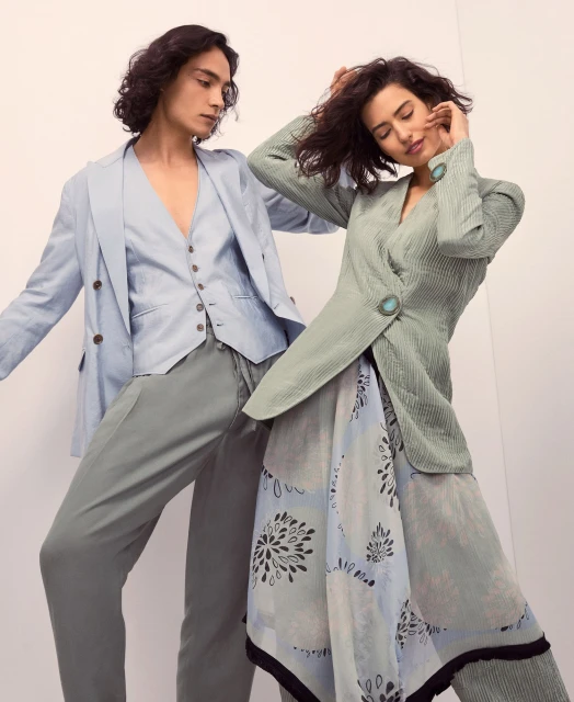 Two fashion models in a studio pose. The female model wears a light green jacket with a print skirt. The male wears a light blue suit.