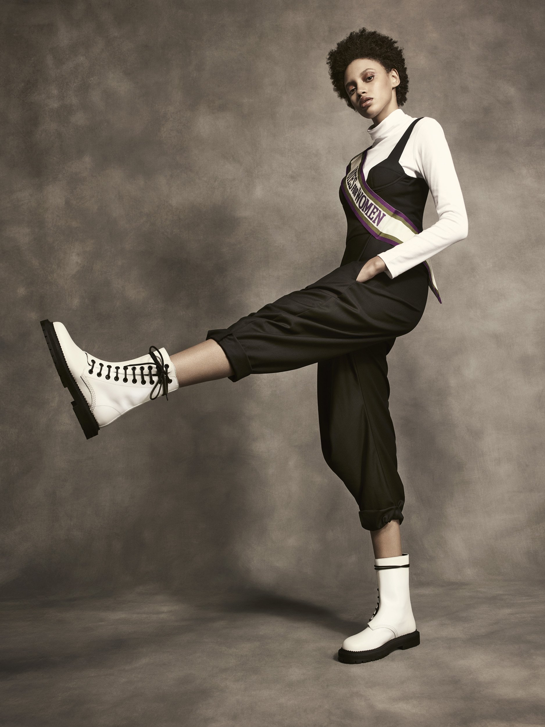 A stylish woman stands in white boots and a black jumpsuit, wearing a sash that reads "VOTE FOR WOMEN," against a grey backdrop.