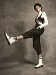 Model with short dark curly hair wearing a white turtleneck, black overalls, sash, and white boots, kicking leg out.