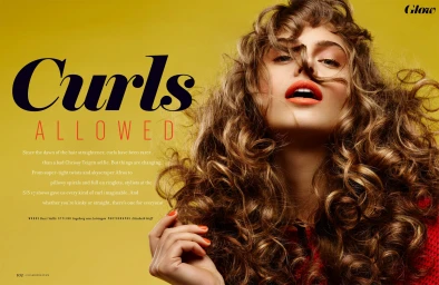 Close-up portrait of a woman with voluminous curly brown hair and bright orange lipstick against a solid yellow background.
