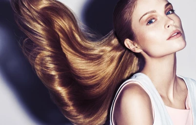 Close-up portrait of a woman with long glossy blonde hair swept in motion against a plain white studio backdrop.
