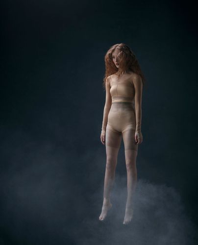 A woman with long, red hair is depicted in a surreal, seemingly levitating pose, set against a gradient dark blue background with subtle smoke effects.