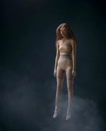 A woman with long, red hair is depicted in a surreal, seemingly levitating pose, set against a gradient dark blue background with subtle smoke effects.