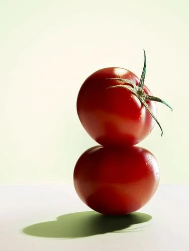 Two vibrant red tomatoes are stacked on a white surface, casting a soft green shadow, against a gradient green background. Food photography.