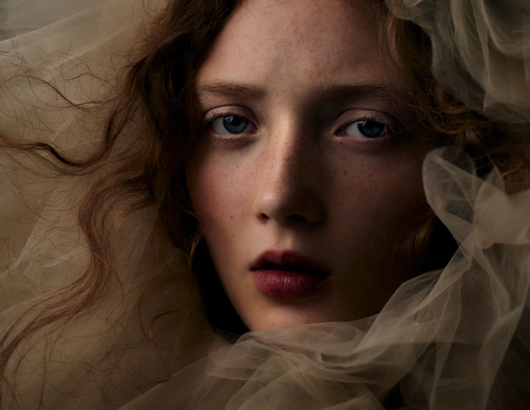 Close-up portrait of a fair-skinned woman with auburn hair, blue eyes, and dark lipstick, surrounded by soft, sheer fabric.