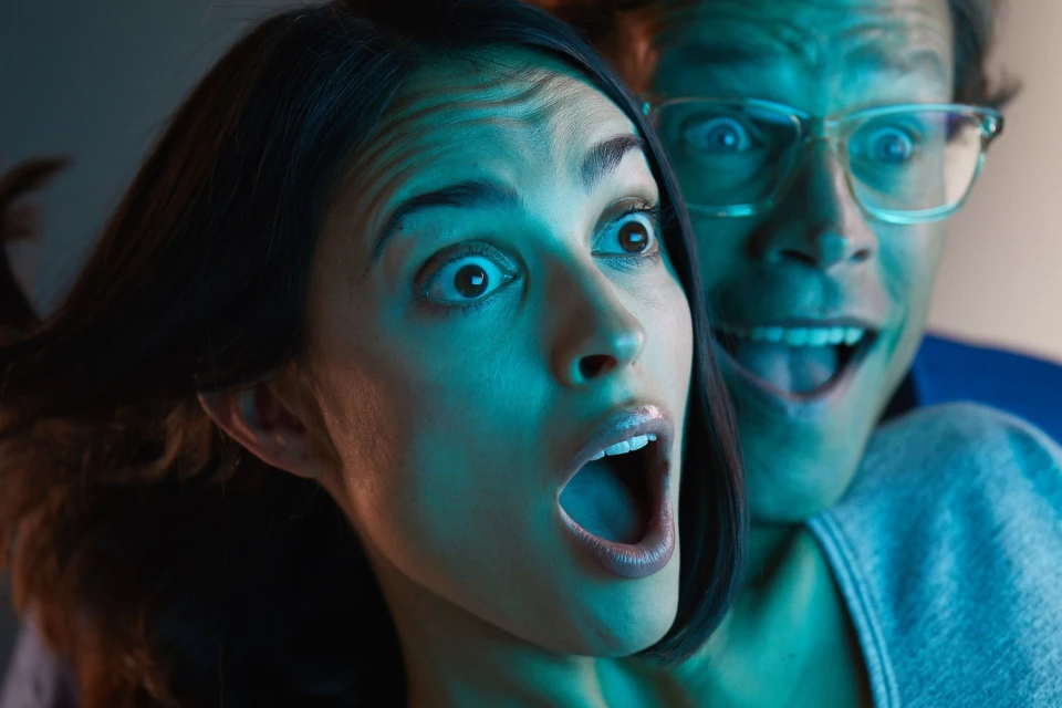 A woman and a man react in fright, with wide eyes and open mouths, bathed in a cool blue light.