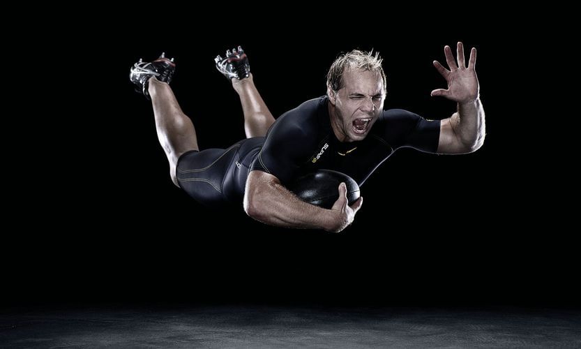 Action shot of a male athlete diving forward and clutching a black rugby ball, captured in a dramatic, high-contrast sports photograph.