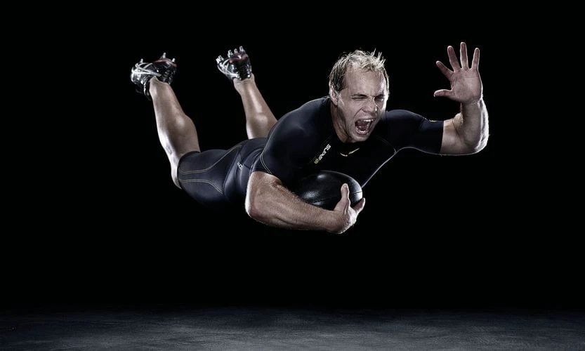 Action shot of a male athlete diving forward and clutching a black rugby ball, captured in a dramatic, high-contrast sports photograph.