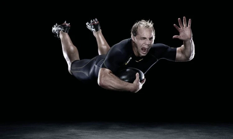 Action shot of a male athlete diving forward and clutching a black rugby ball, captured in a dramatic, high-contrast sports photograph.