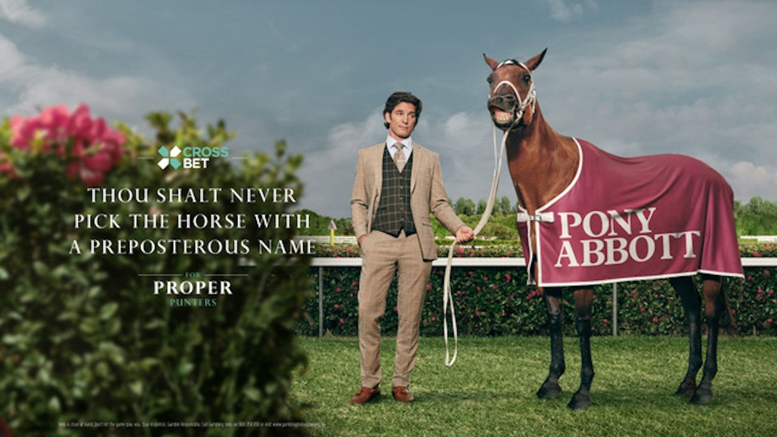 A man in a suit stands next to a horse wearing a blanket that says Pony Abbott, in an outdoor setting with greenery and signage.
