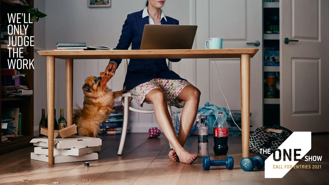 Woman in business attire works at a table with a laptop; she wears pajama shorts and a dog tries to grab pizza from her. 