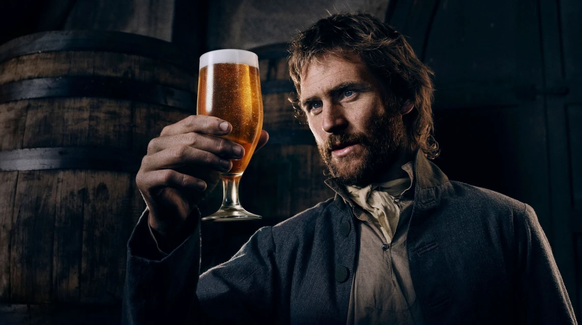 Bearded man in historical clothing holding glass of beer in a dark cellar with wooden barrels, moody lighting, and rustic charm.