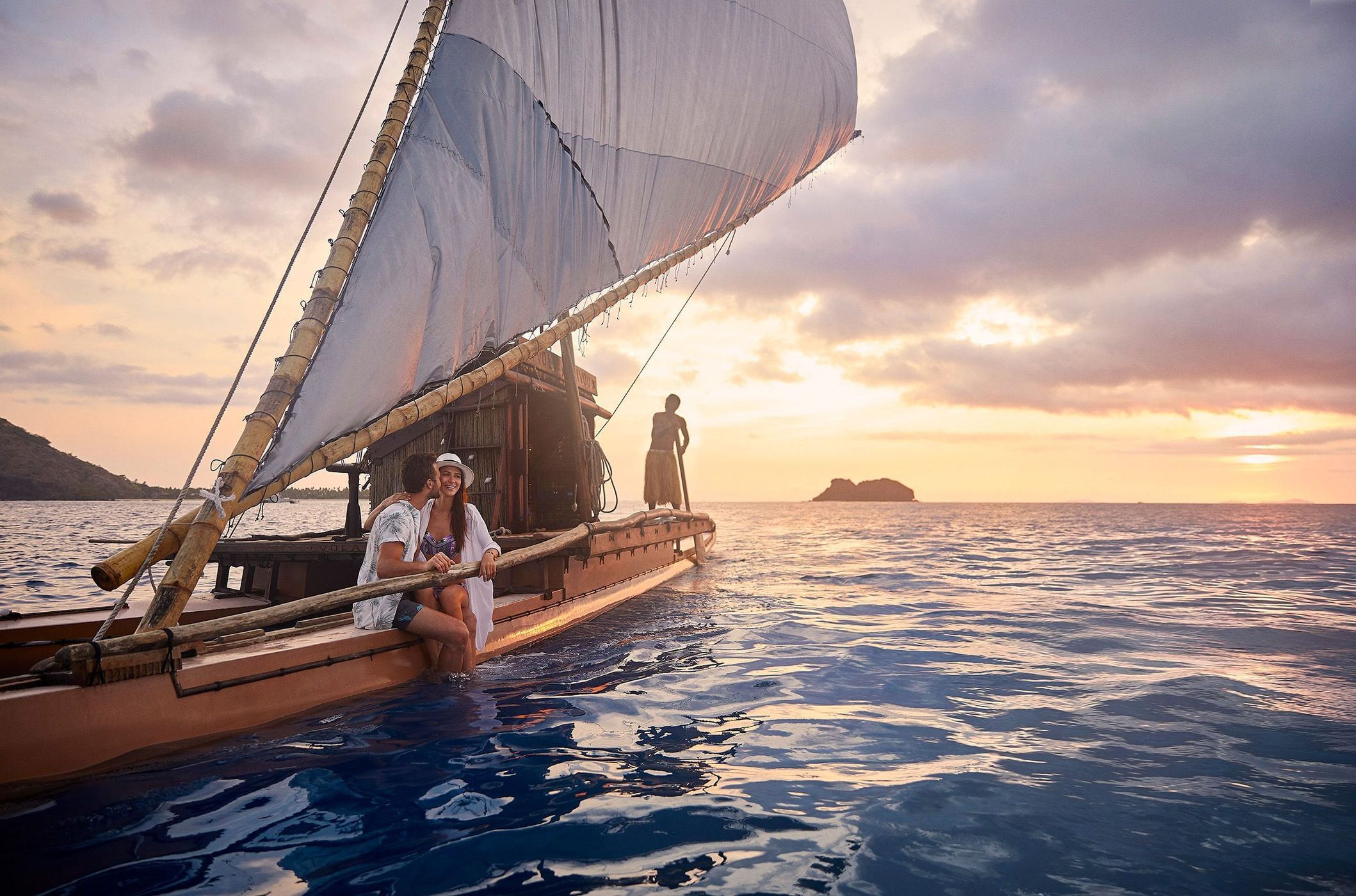 Couple enjoys a boat ride at sunset on a traditional outrigger sailing vessel, with a paddler silhouetted against the warm sky.