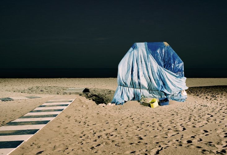 Beach scene with striped walkway leading to a blue-covered structure, buckets nearby, under a dark sky and ocean horizon.