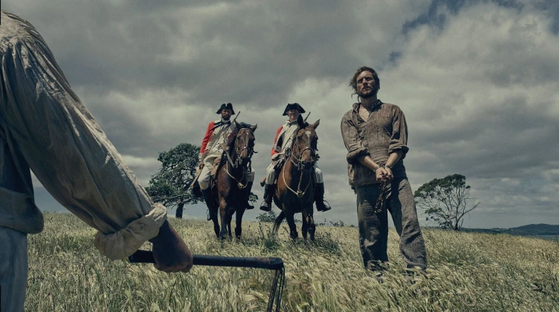 Man in shackles stands in a field, guarded by two mounted soldiers in red coats, under an overcast sky, suggesting a historical drama.