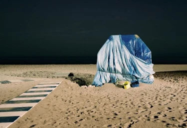 Covered structure wrapped in blue tarp on a sandy beach at night under a dark sky