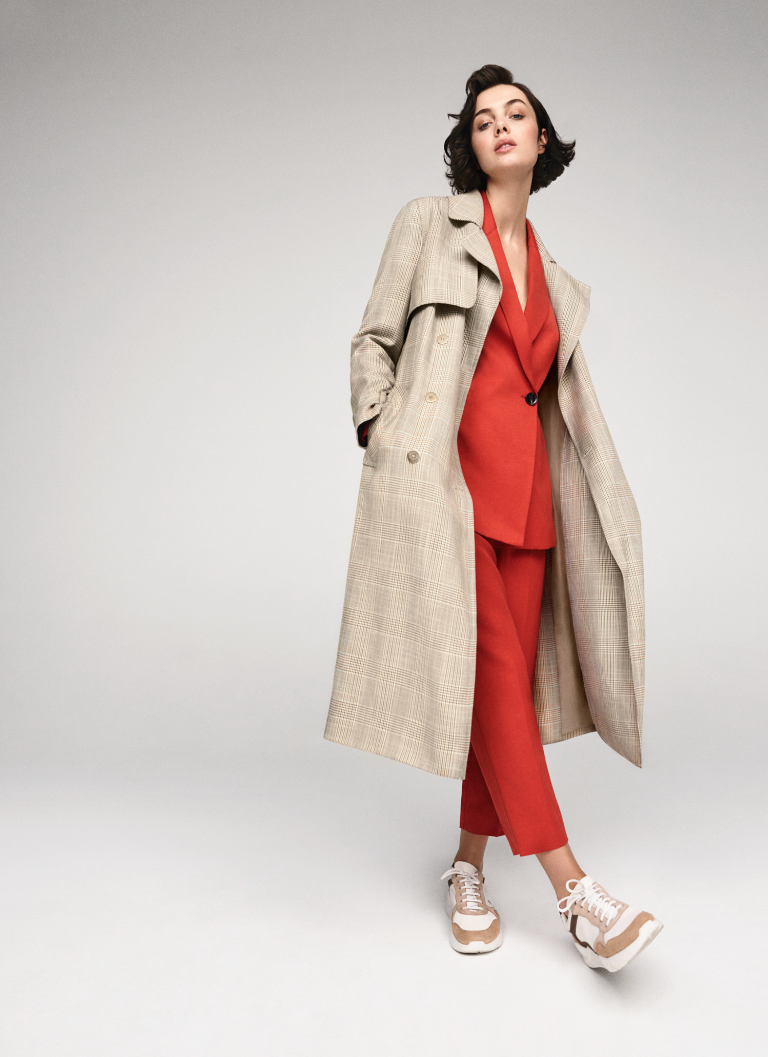 Fashionable woman in a red pantsuit and plaid trench coat stands in a neutral studio, hands in pockets, wearing beige sneakers.