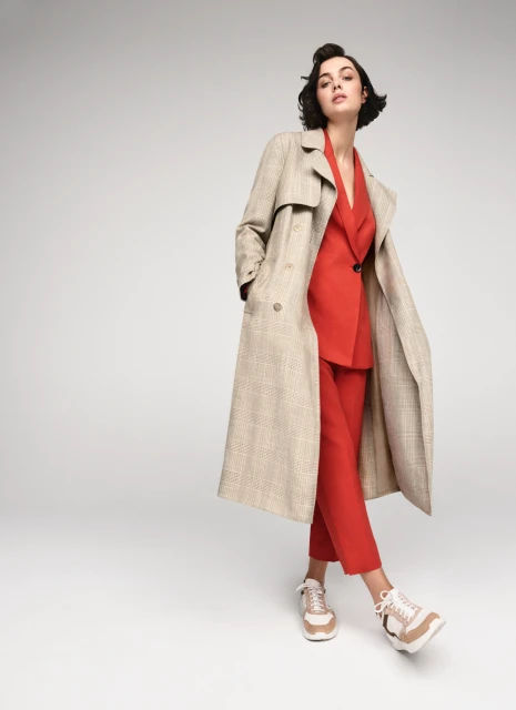 Fashionable woman in a red pantsuit and plaid trench coat stands in a neutral studio, hands in pockets, wearing beige sneakers.