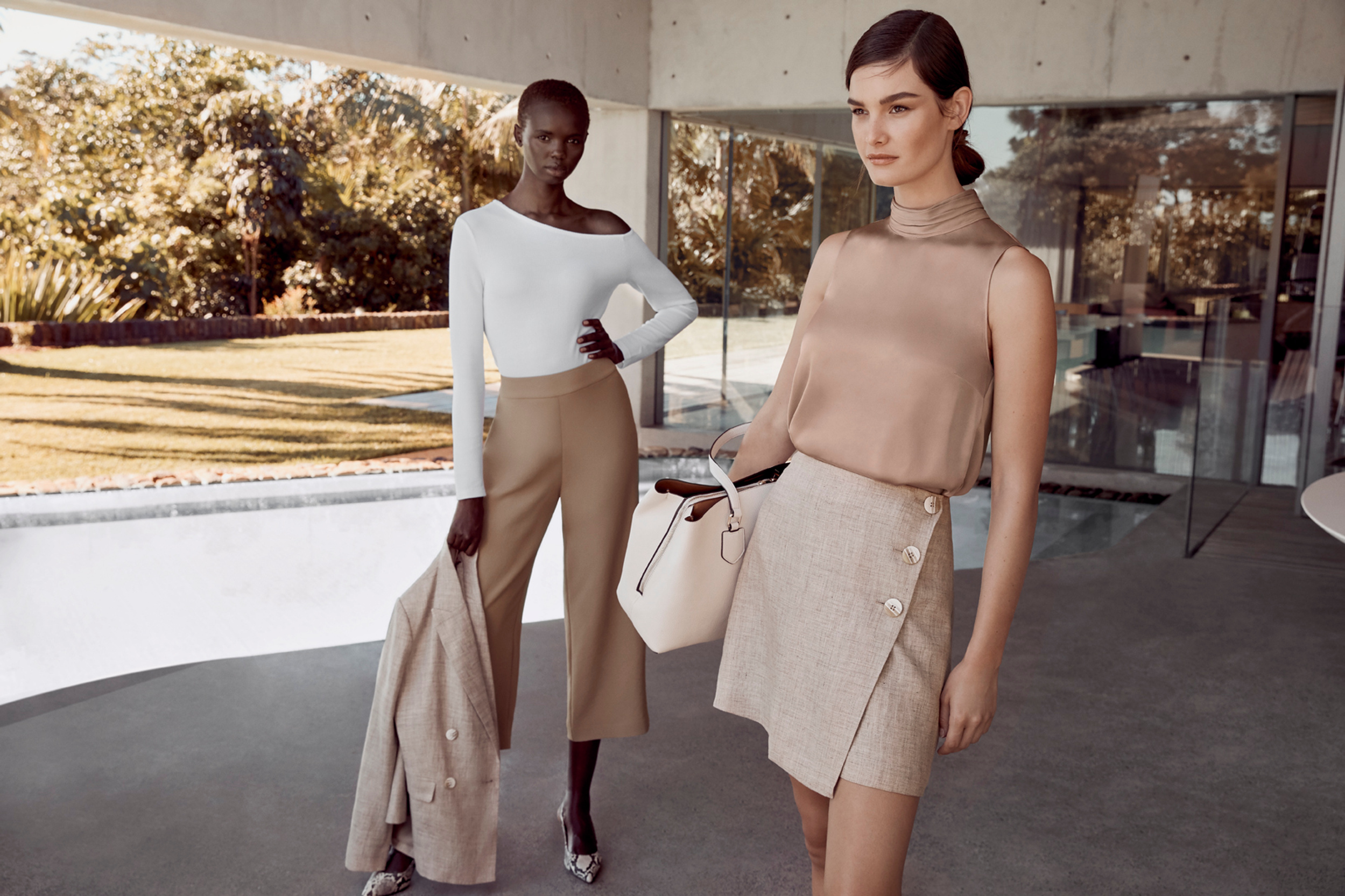 Two fashion models in stylish, neutral-tone professional attire pose near a modern building with a lawn and large windows.