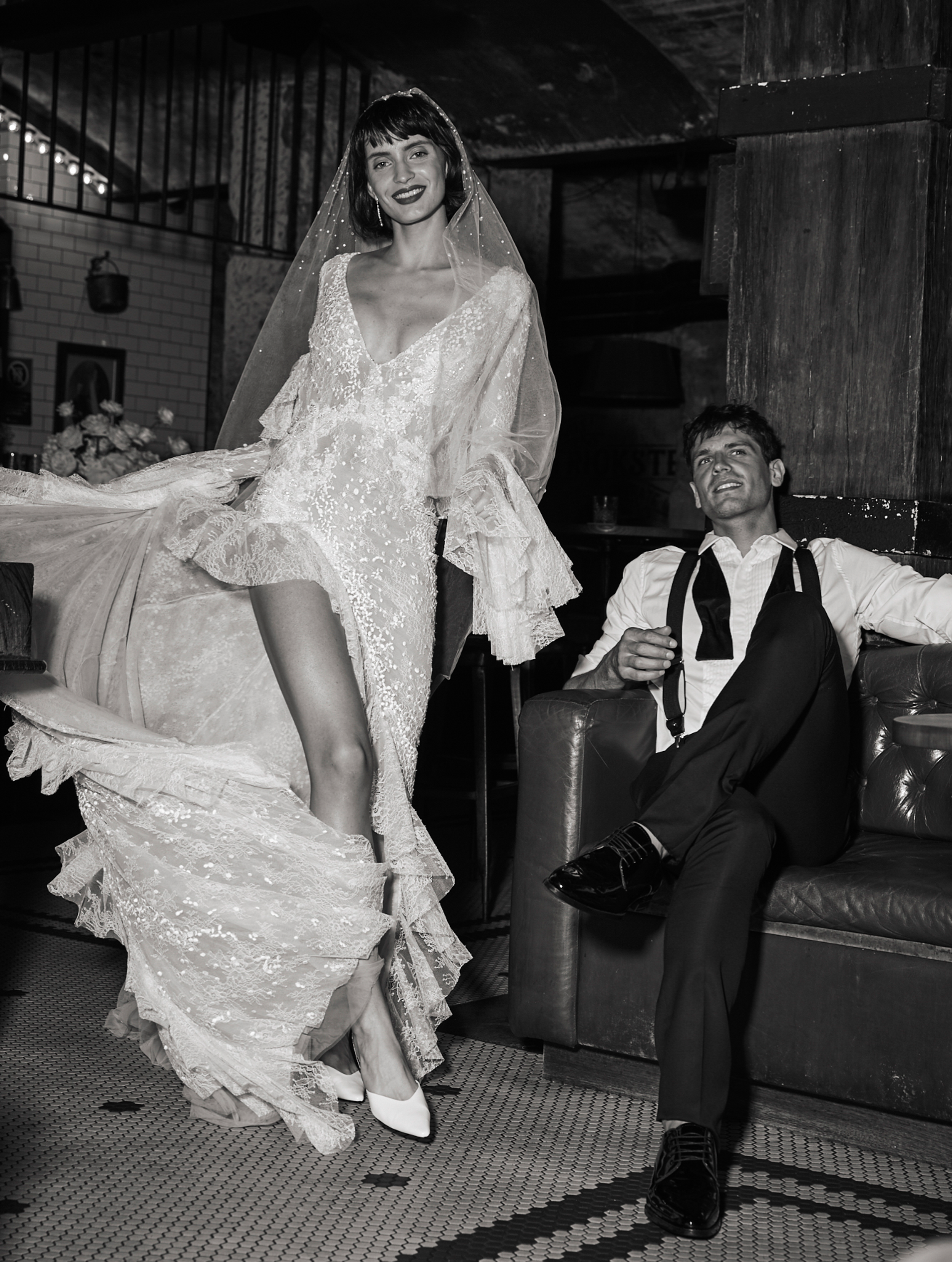 Bride in lace wedding dress with groom in suit on couch, dark interior setting, black-and-white fashion shot.