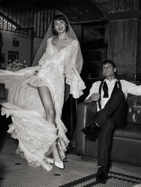 Bride in lace wedding dress with groom in suit on couch, dark interior setting, black-and-white fashion shot.